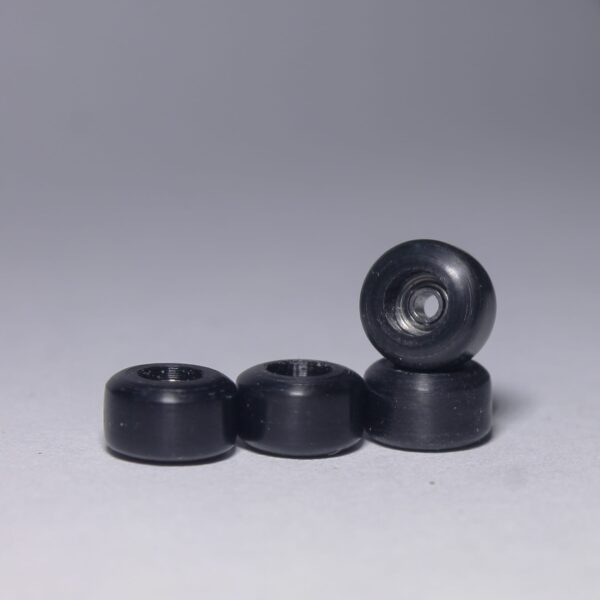 3 black fingerboard wheels laying on their back side. The fourth wheel is standing up right on the opening of the laying on its side furthest to the right.